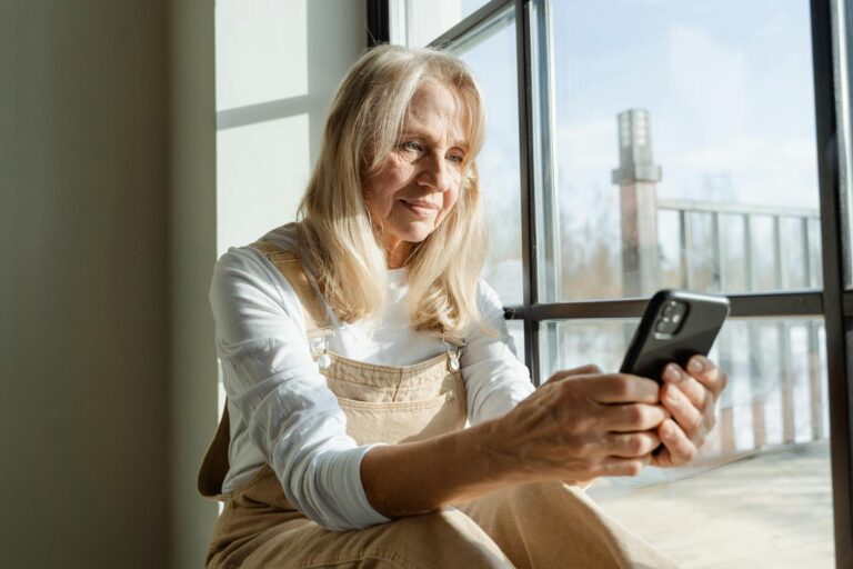 Elderly woman using a smartphone by a window, enjoying leisure time indoors.