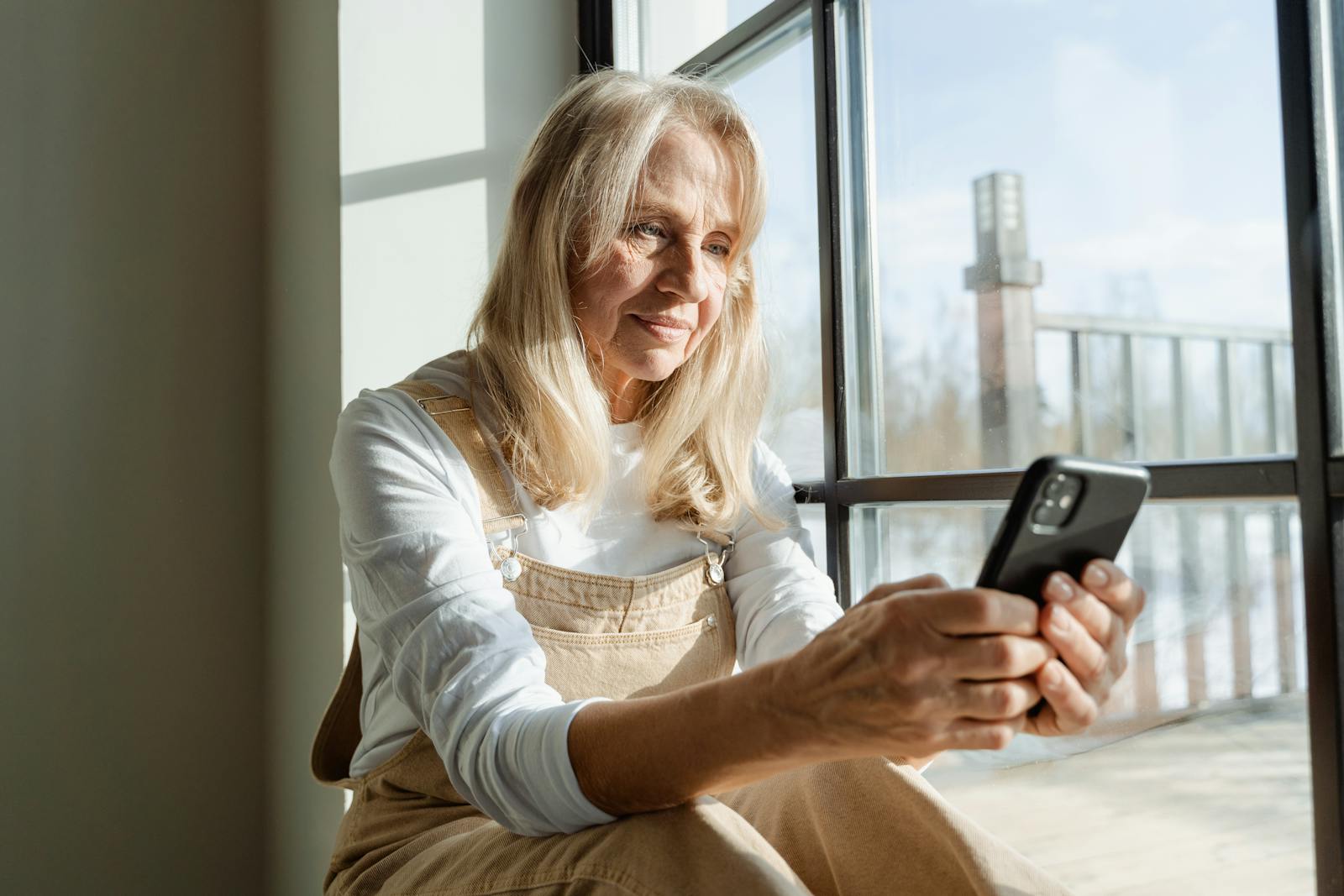 Elderly woman using a smartphone by a window, enjoying leisure time indoors.