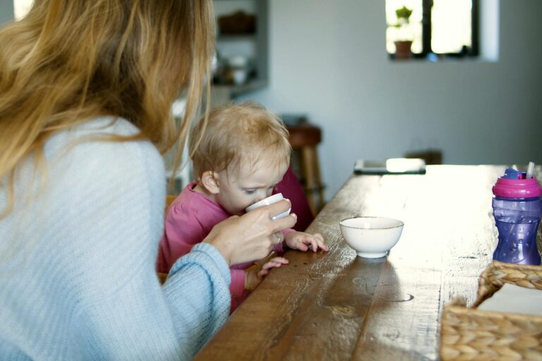 A mother helps her child drink from a cup on a wooden table in a cozy home setting.
