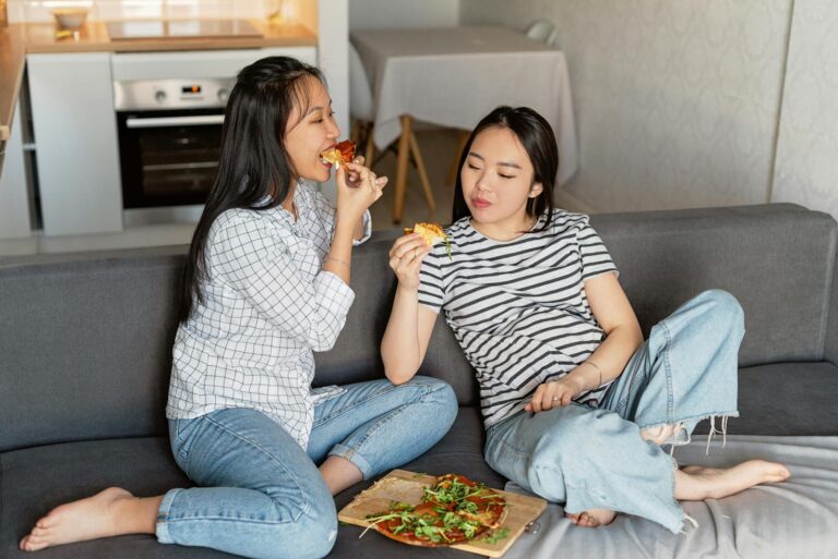 Two women sitting on a sofa at home, enjoying slices of pizza, embracing leisure time.
