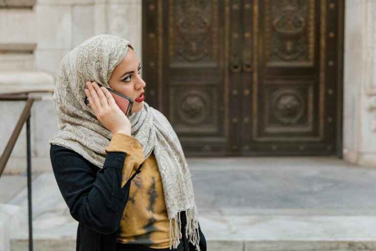 Middle Eastern woman wearing hijab talks on phone in urban setting.