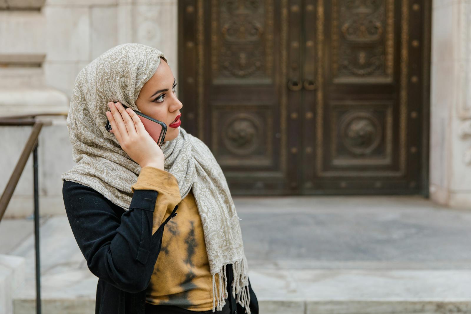 Middle Eastern woman wearing hijab talks on phone in urban setting.
