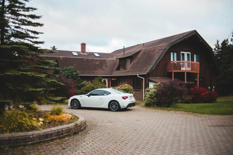 Rustic house with a white car parked on a cobblestone driveway amidst lush greenery.