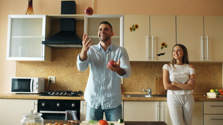 Man juggles apples while woman watches in kitchen.