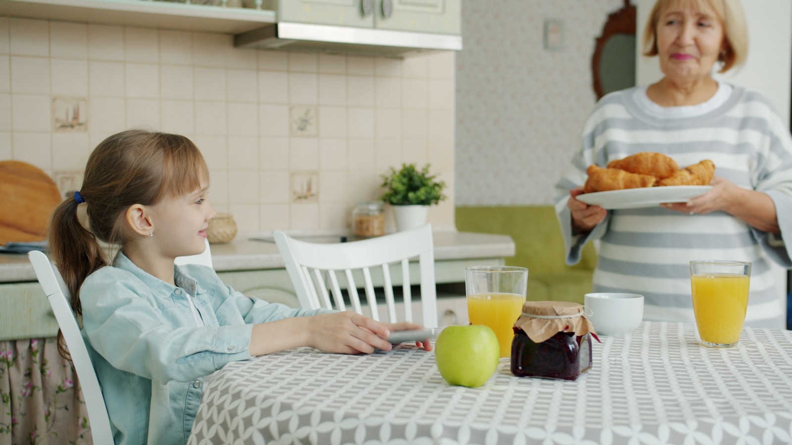 Grandmother serves croissants to girl at kitchen table.