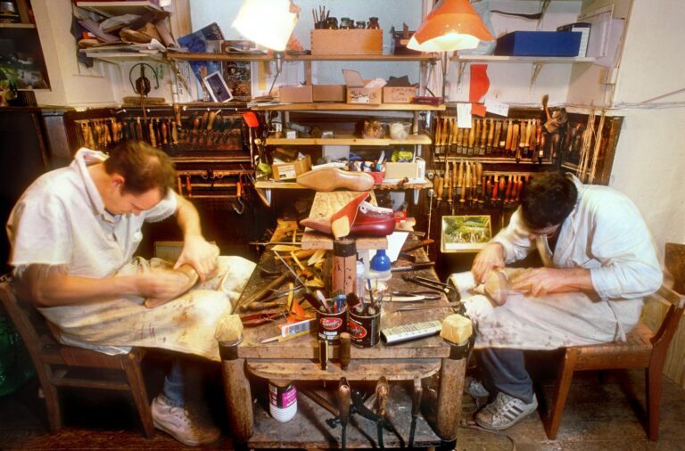 Two men crafting shoes in a cluttered workshop filled with tools and materials.
