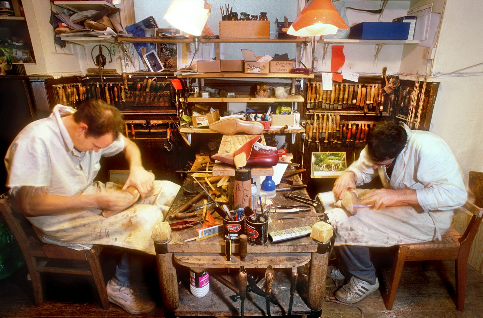 Two men crafting shoes in a cluttered workshop filled with tools and materials.