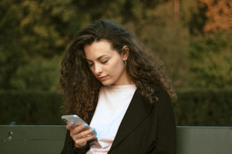 Woman with curly hair checking smartphone while sitting outdoors in Warsaw park.
