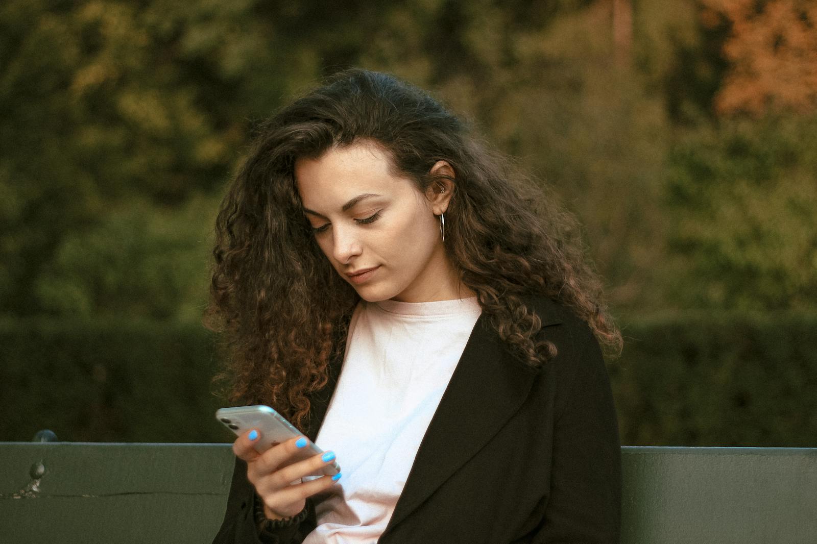 Woman with curly hair checking smartphone while sitting outdoors in Warsaw park.