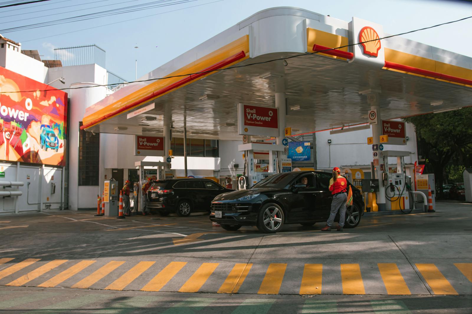 A Shell gas station with vehicles refueling and attendants assisting drivers on a sunny day.