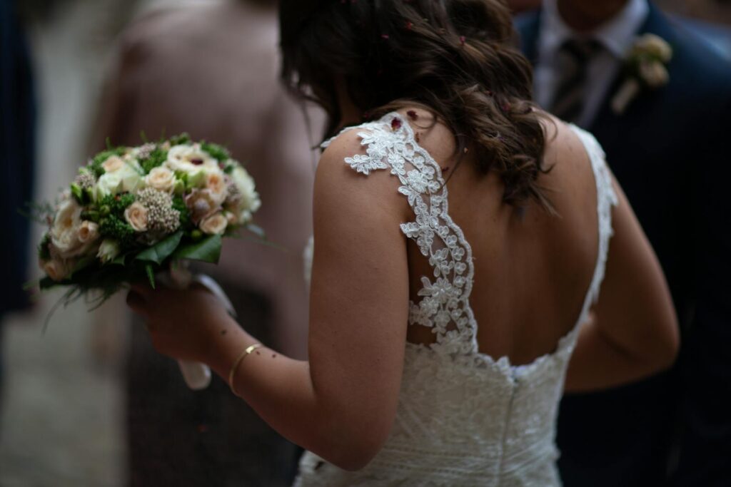 A bride in a lace gown holds a beautiful bouquet, viewed from behind at a wedding ceremony.