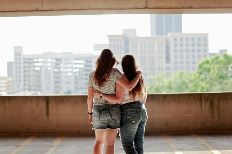 Two women in casual clothing embrace with a city skyline in the background.