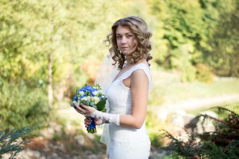 Beautiful bride in white dress holding blue bouquet outdoors in a sunny park.