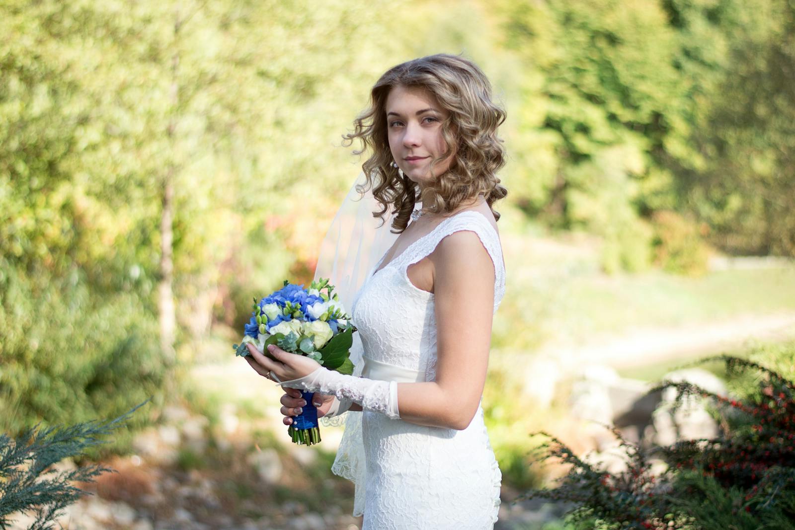 Beautiful bride in white dress holding blue bouquet outdoors in a sunny park.