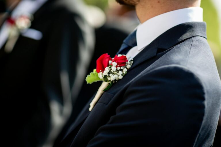 Close-up of a stylish man in a suit adorned with a vibrant rose boutonniere at a daytime wedding.