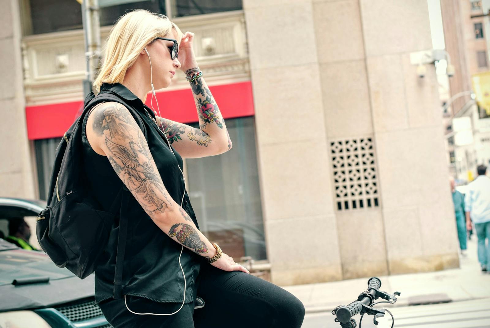 Woman with tattoos listens to music while resting on a bike in city setting.