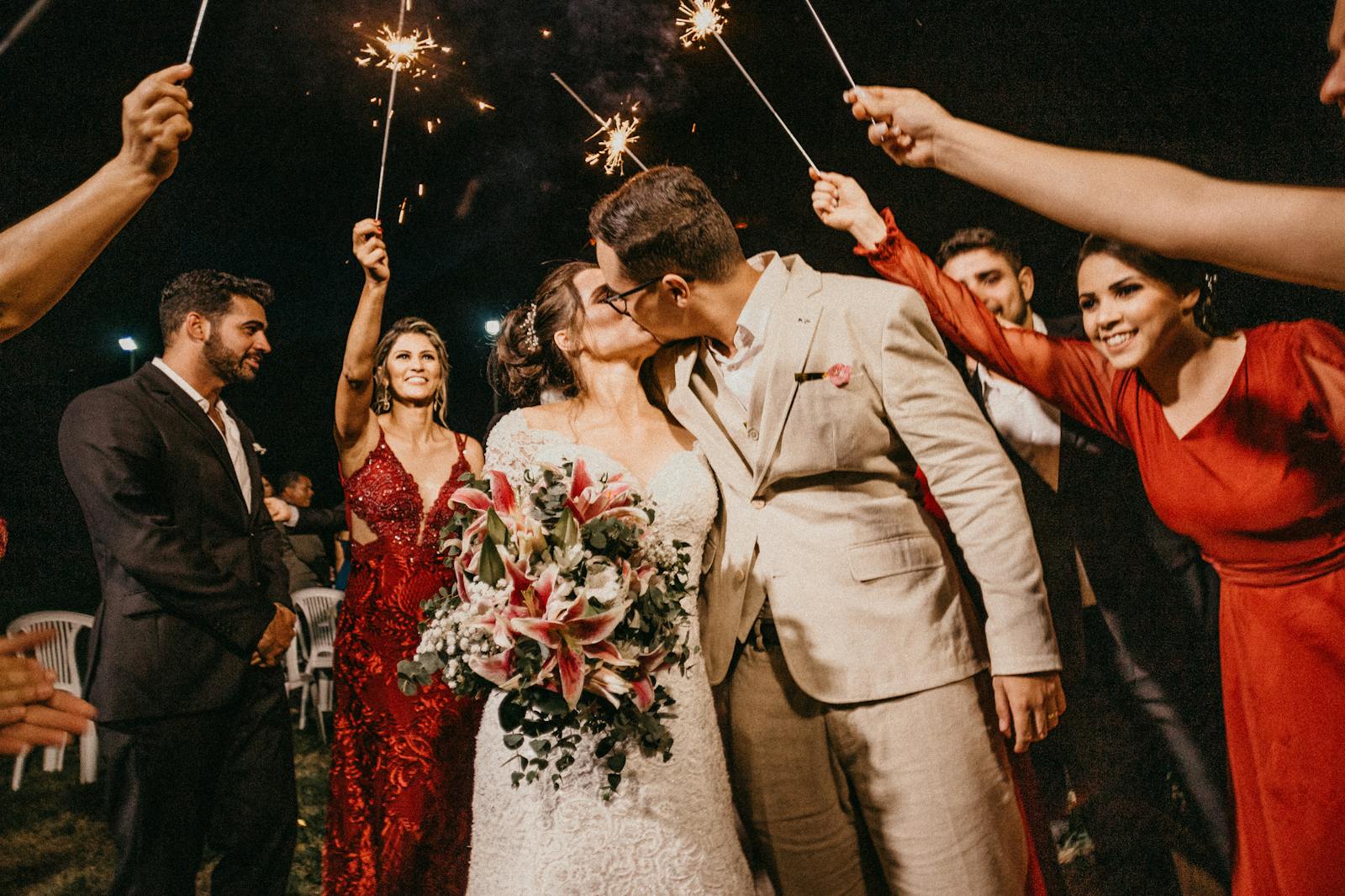 A joyful couple kisses under sparklers, surrounded by guests in a wedding celebration.