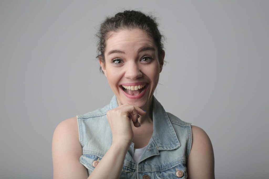 Portrait of a joyful young woman with a big smile, wearing a casual denim vest indoors.