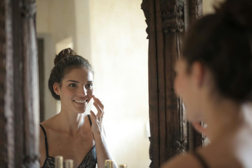 Young woman applying skincare and smiling at her reflection in a rustic mirror.