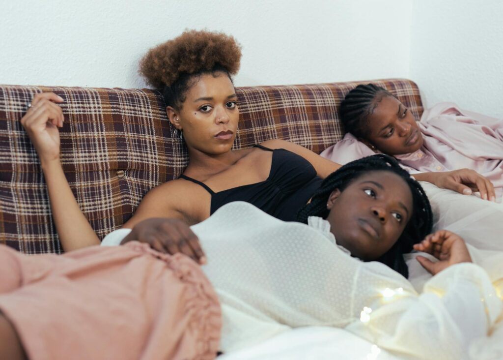 Three black women relaxing on a bed in a cozy indoor setting, reflecting a moment of calm and companionship.