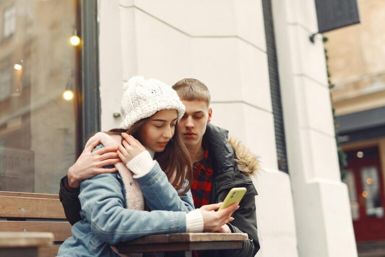 A loving couple sits together outside, sharing a cozy winter moment with a smartphone.