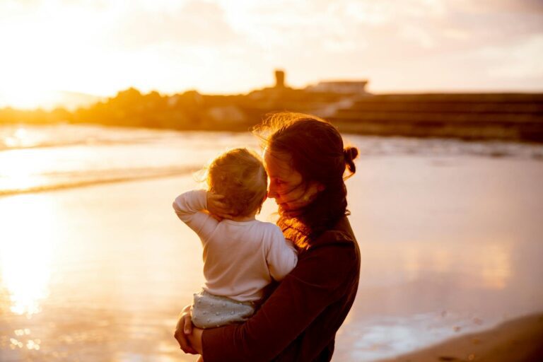 A loving mother embraces her child on a sunlit beach during sunset, celebrating family and togetherness.