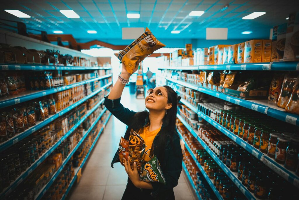 A woman gleefully holds bags of potato chips in a supermarket aisle, capturing a moment of joy while shopping.