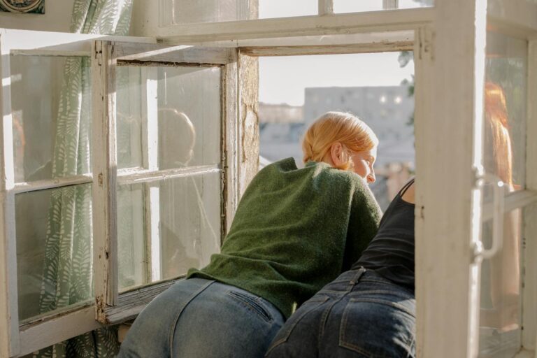 Two women enjoying a sunny view from an open window, creating a serene indoor scene.