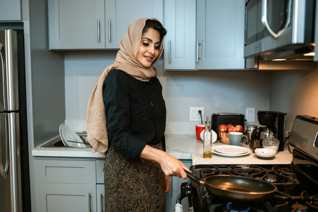 A cheerful woman wearing a headscarf cooks a meal in a contemporary kitchen.