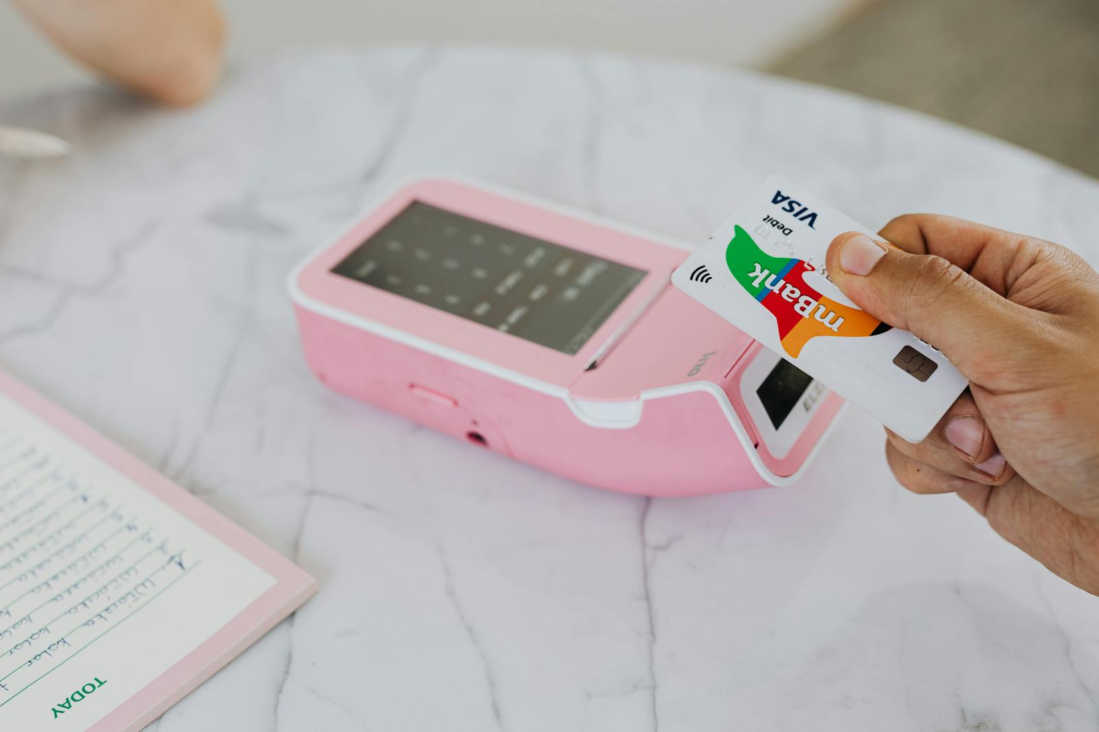 A hand using a Visa card for a contactless payment on a pink terminal.
