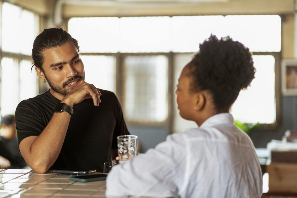 Two adults conversing at a table indoors, with natural light through windows.