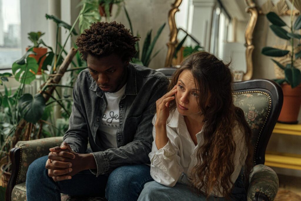 A couple sits together on a couch surrounded by indoor plants, deep in thought.