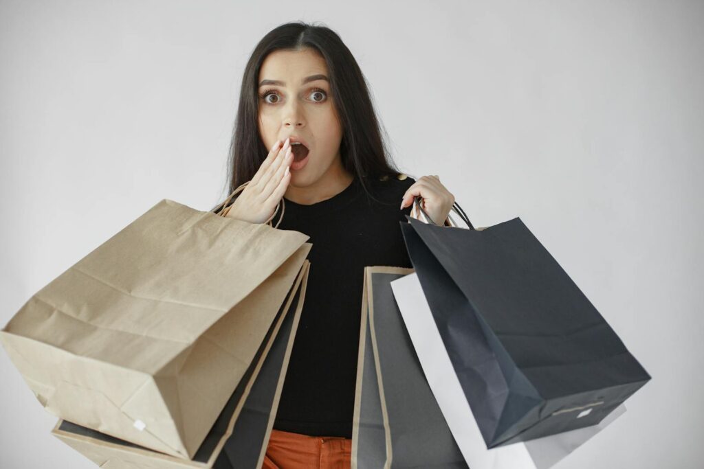 Woman shocked with shopping haul, holding multiple paper bags indoors.