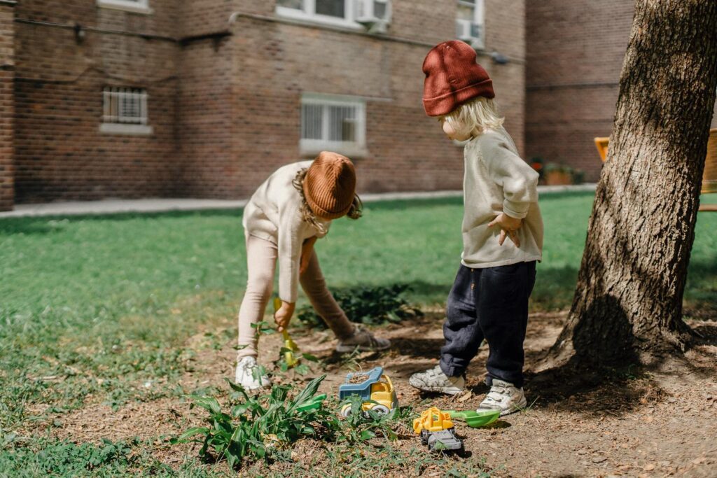 Unrecognizable little kids in casual outfits and brown hats playing with toys on backyard beside big tree