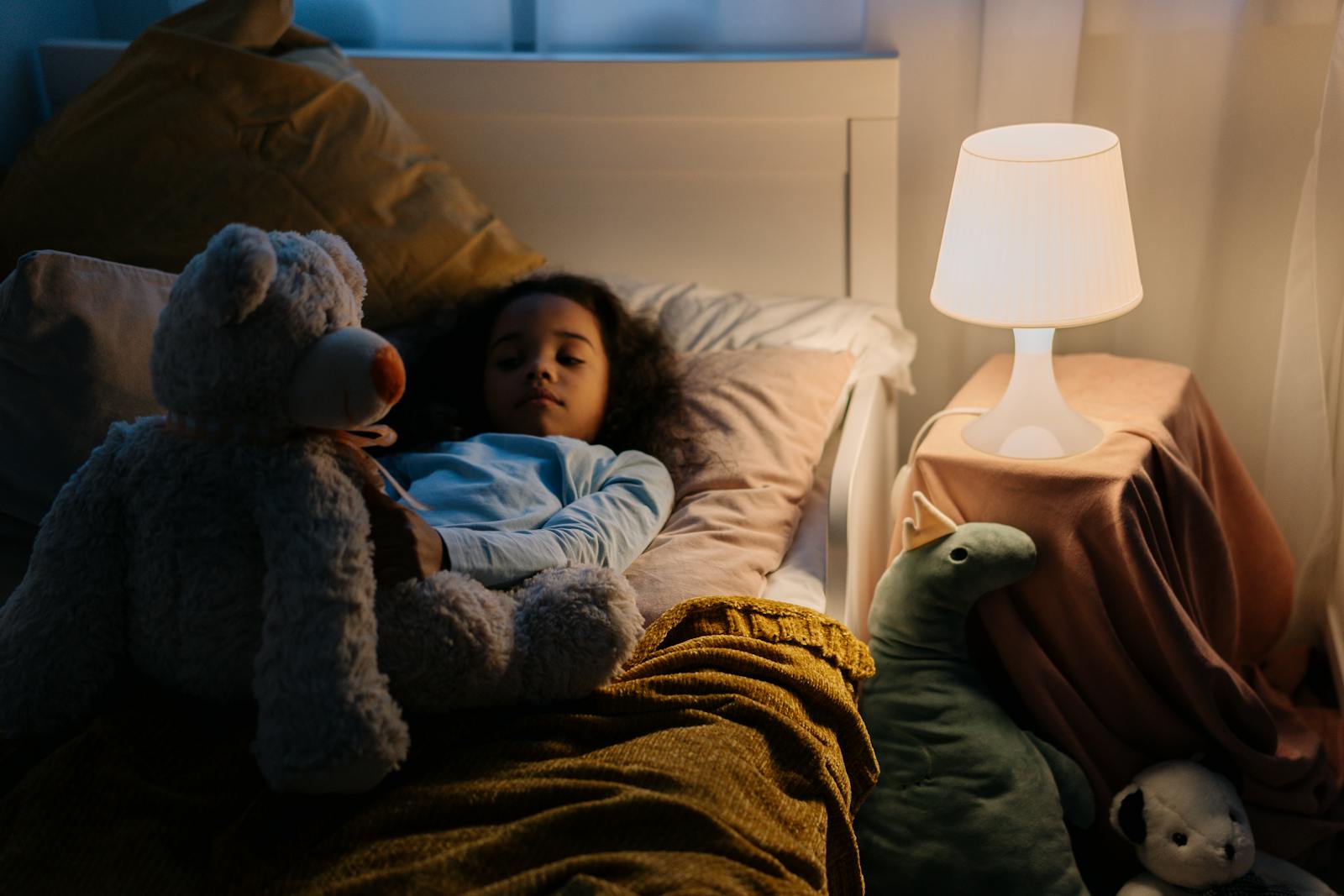 Young girl resting in bed with a teddy bear and toys, illuminated by a bedside lamp.