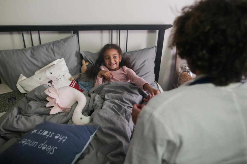 A smiling child lying in bed surrounded by plush toys in an indoor bedroom setting.