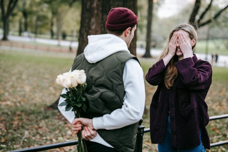 Young couple enjoying a romantic surprise moment in a sunny park, with hidden flowers and a vibrant autumn backdrop.