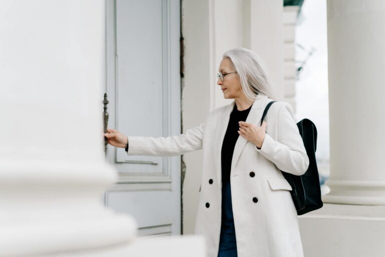 Stylish senior woman in white coat opening a door while standing outdoors, exuding confidence and grace.