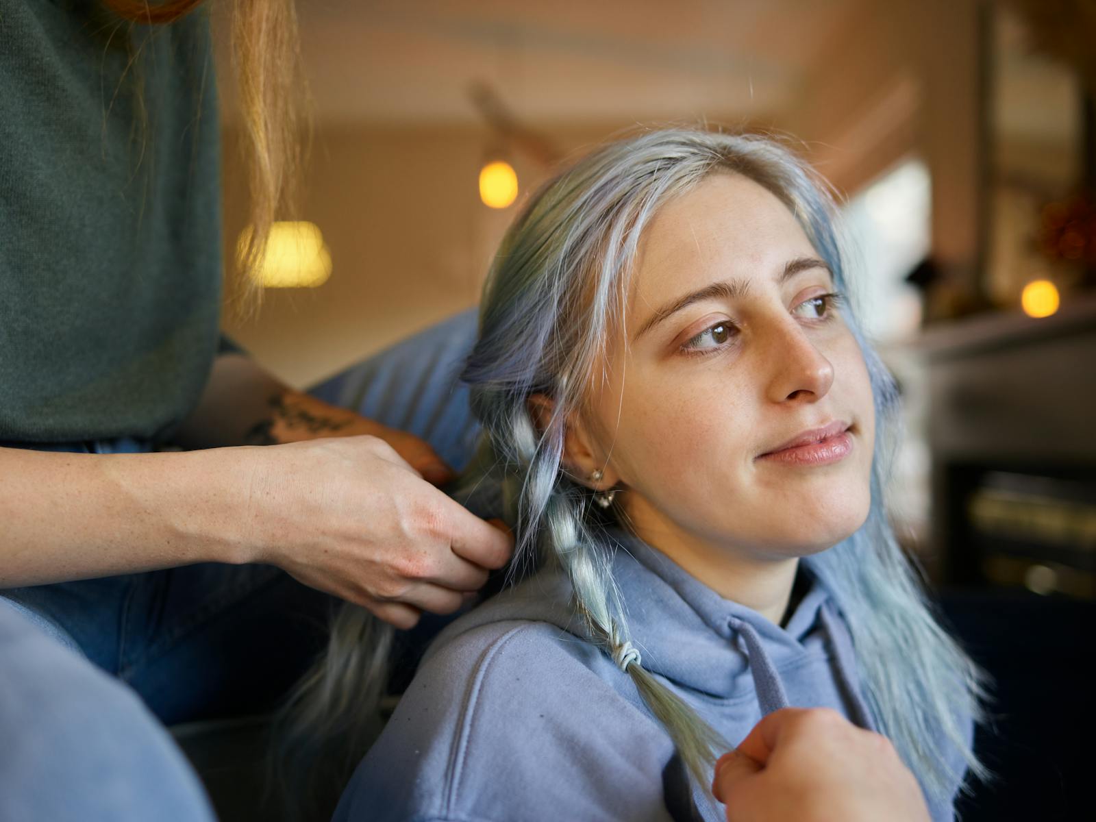 A young woman with gray hair getting a braid done indoors with a gentle smile.