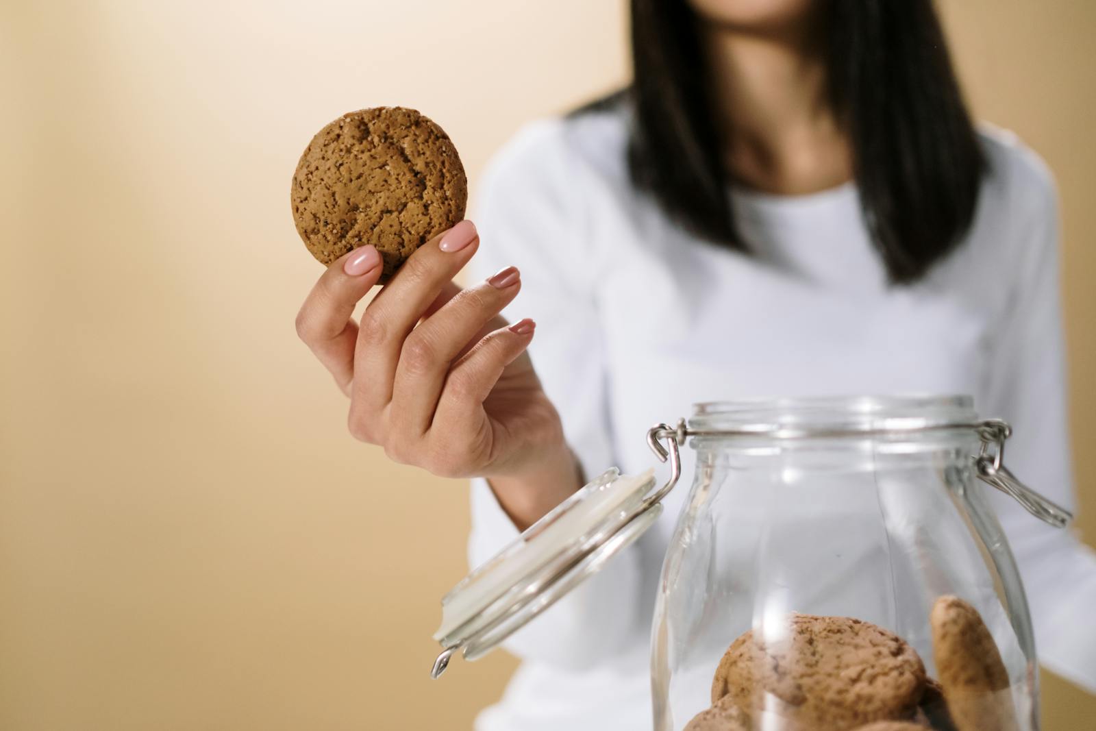 Close-up of a woman's hand holding a cookie from a glass jar, with a soft focus background.