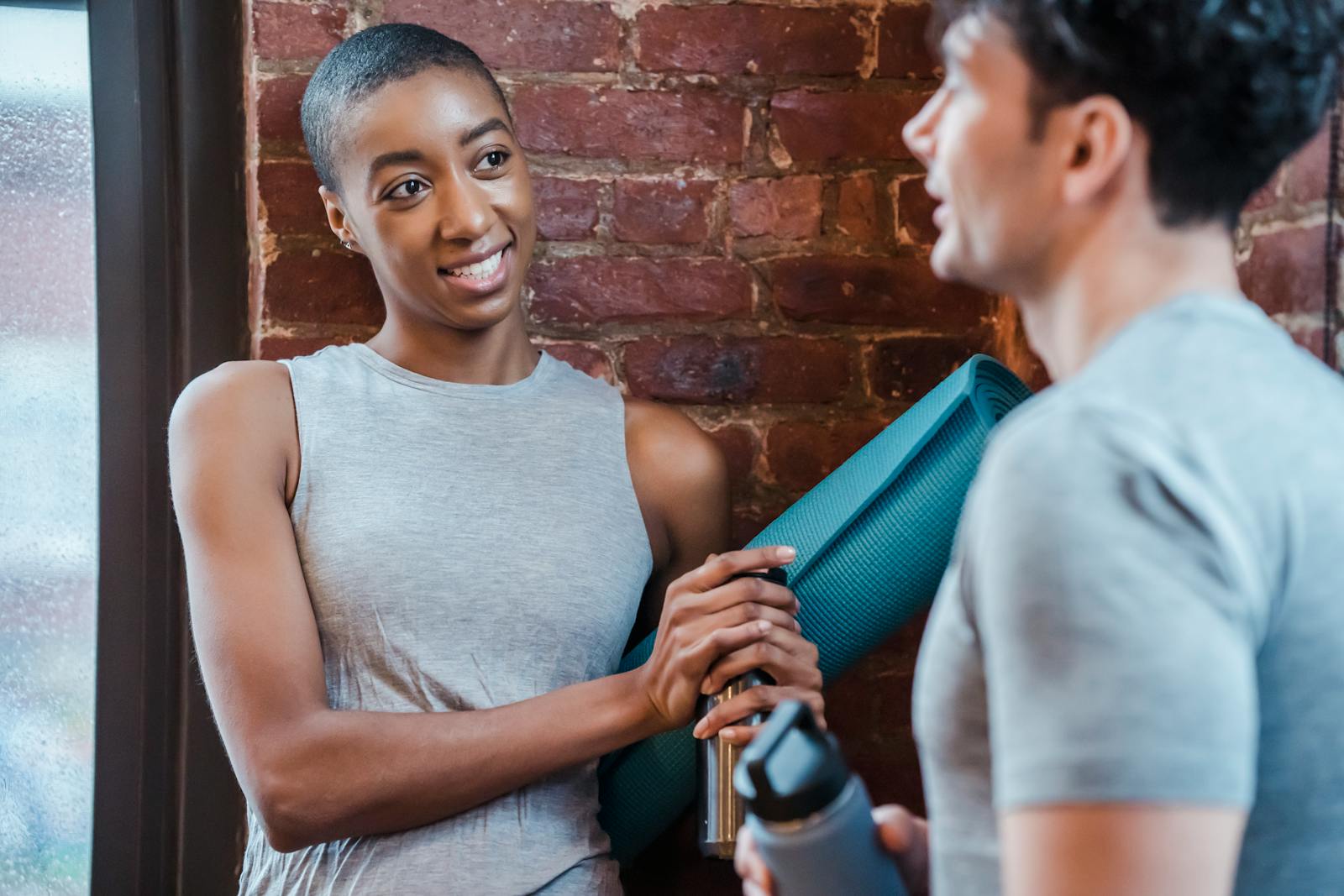 Two diverse friends engaging in conversation after a workout, holding yoga mats and water bottles.