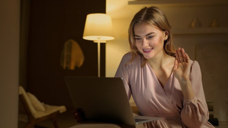 Woman smiling and waving during a video call, using a laptop in a cozy indoor setting.