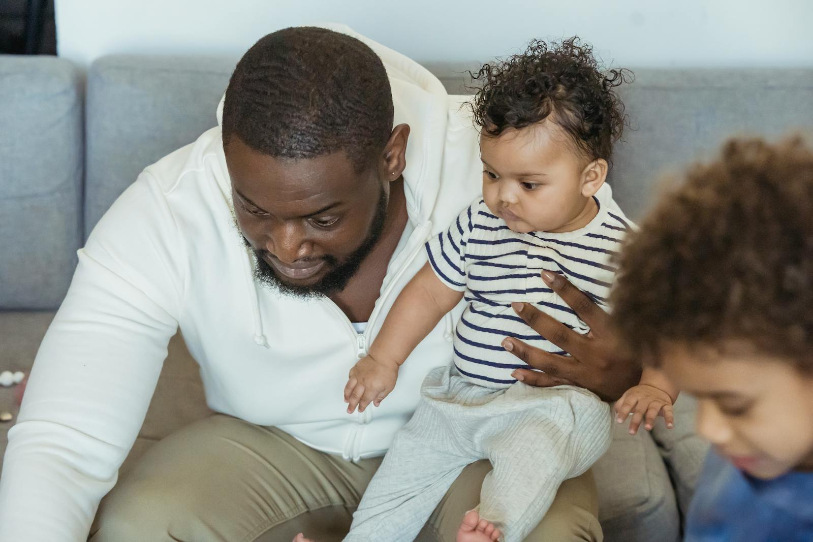 Bearded African American dad with cute little toddler in arms near crop son in lounge