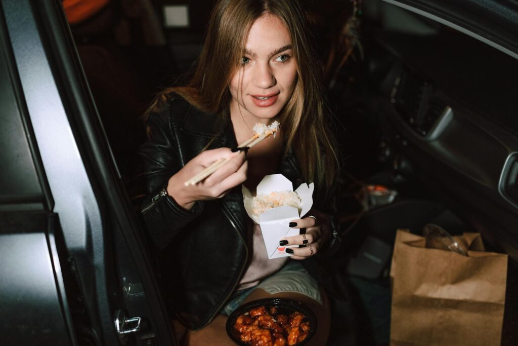Young woman eating Asian takeaway food in car, using chopsticks and seated in black leather jacket.