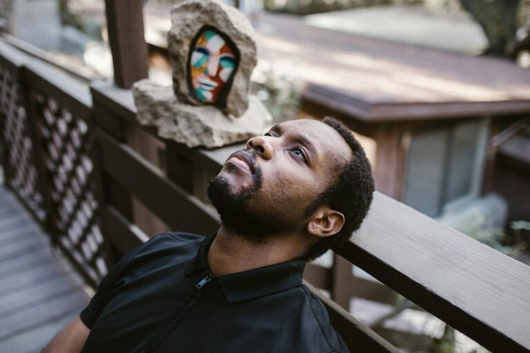 A man in thoughtful reflection sits outdoors against a wooden railing with abstract art behind.