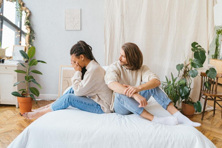 Couple sitting back-to-back on bed, showing relationship conflict in a cozy room.