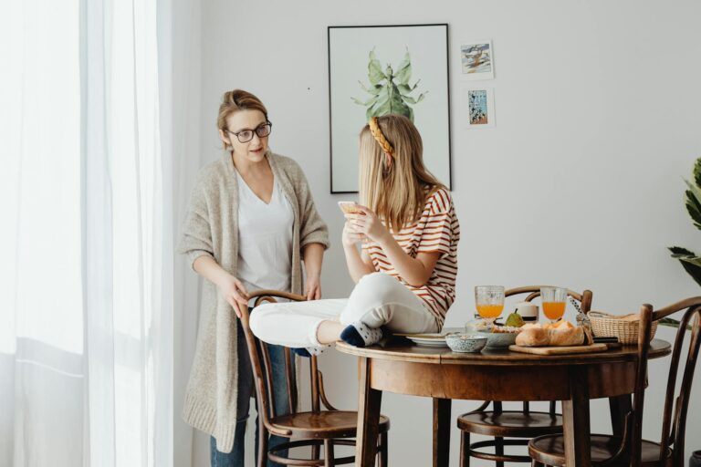 A mother and daughter share a moment at the dining table in a warm, domestic setting with breakfast items.