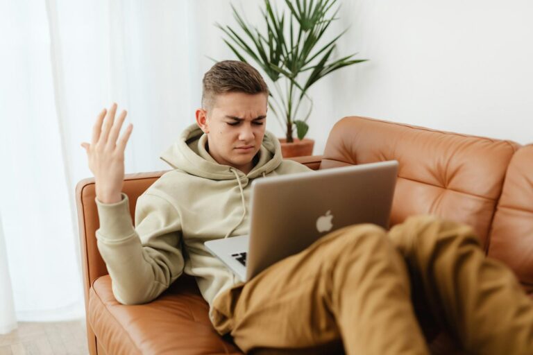 A young man in a hoodie looks irritated while working on a laptop in a cozy indoor setting.