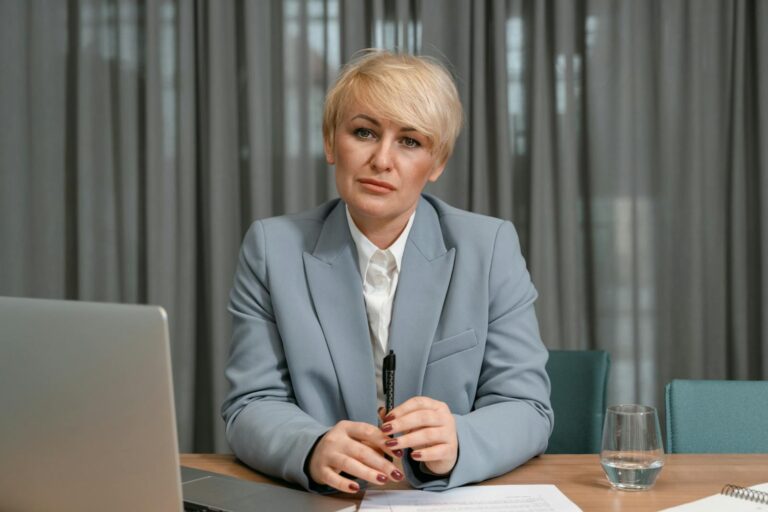 A confident businesswoman in a blue suit sits at her desk with a laptop, pen, and papers in an office setting.