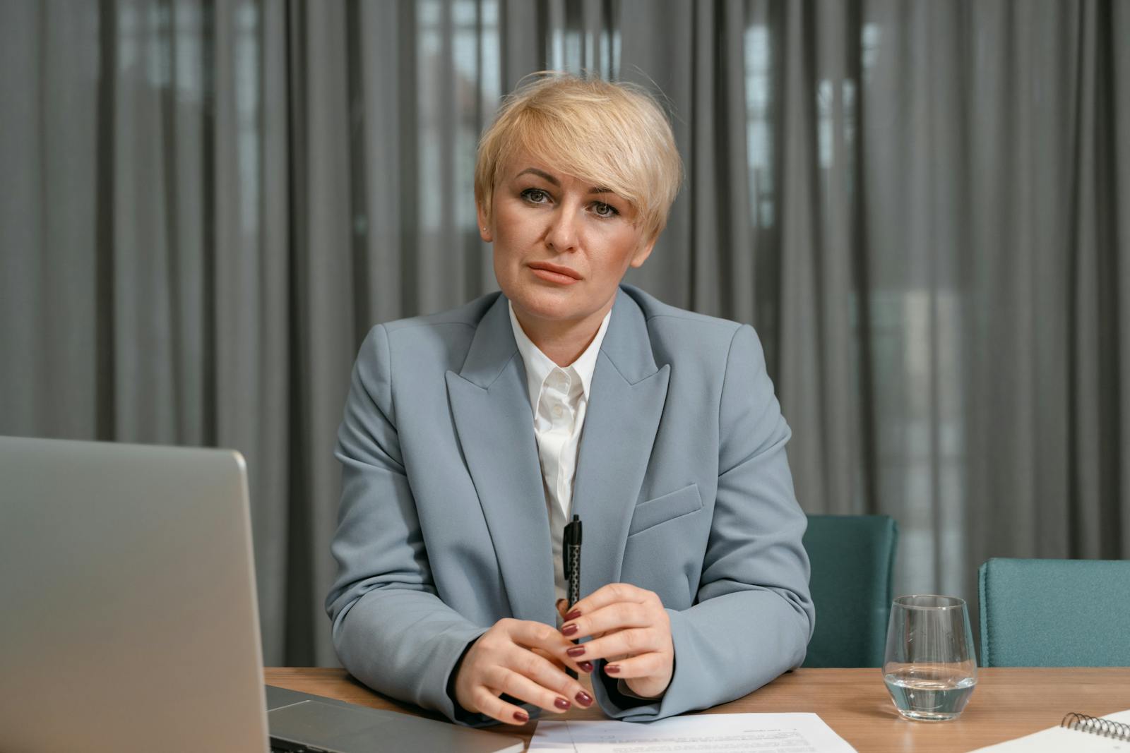 A confident businesswoman in a blue suit sits at her desk with a laptop, pen, and papers in an office setting.